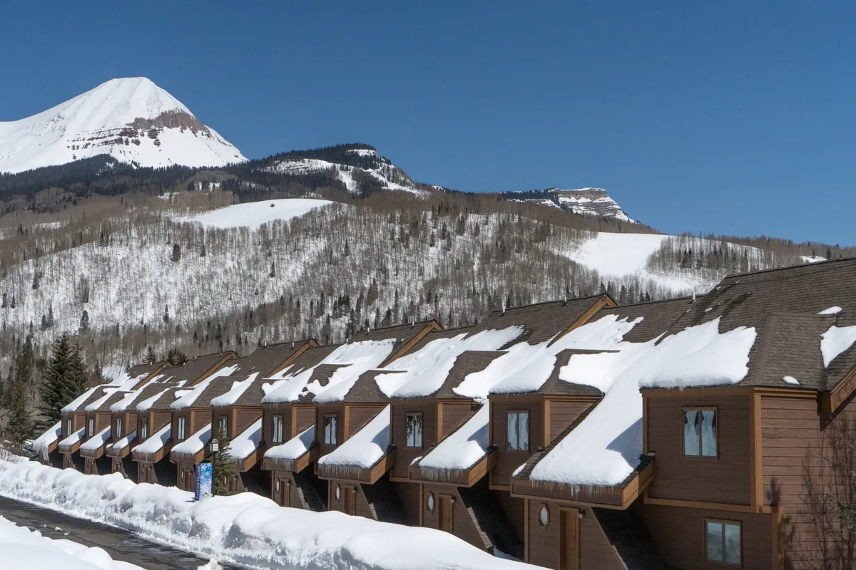 An exterior scene showing snow-covered lodge rooftops against a dramatic mountain backdrop with clear blue skies, encapsulating the cozy, welcoming mood of the resort setting. Subtle hints of ski activity and the inviting lodge exterior connect to the full Purgatory experience.