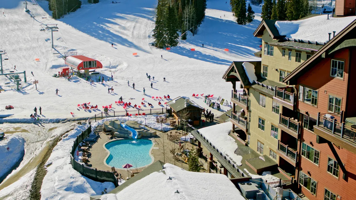 This vibrant exterior image centers on the outdoor pool area at the base of the ski slopes, with people enjoying both swimming and relaxing poolside while surrounded by lodge buildings and dramatic mountain views. The energy of active skiers and the nearby ski lift contribute to the lively, resort-style atmosphere emphasized in the section.