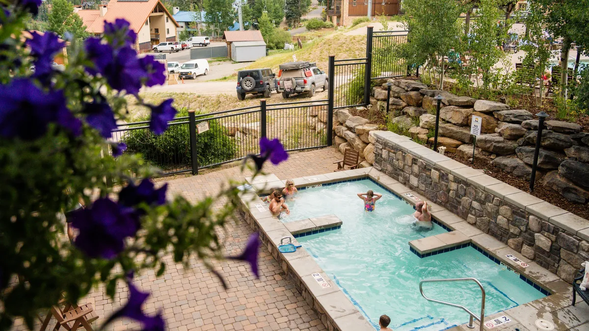 An inviting outdoor pool surrounded by lush landscaping, with people swimming and relaxing in nearby hot tubs. The scene is vibrant, with guests lounging, socializing, and enjoying sunny weather.