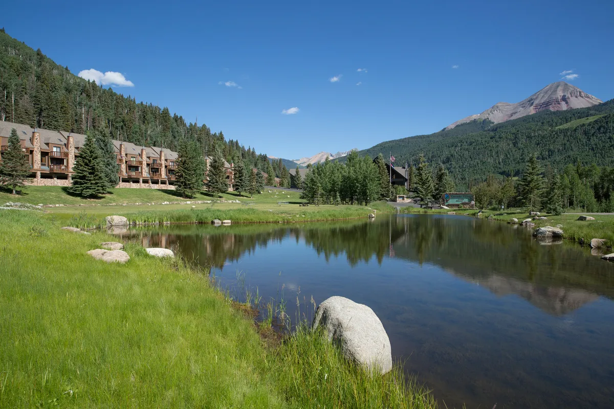 A serene pond reflecting a stunning mountain backdrop, surrounded by lush greenery and wooden buildings. The setting is tranquil yet inviting, suggesting a range of outdoor activities such as fishing, picnicking, and wildlife watching—perfect for families seeking memorable experiences in nature.