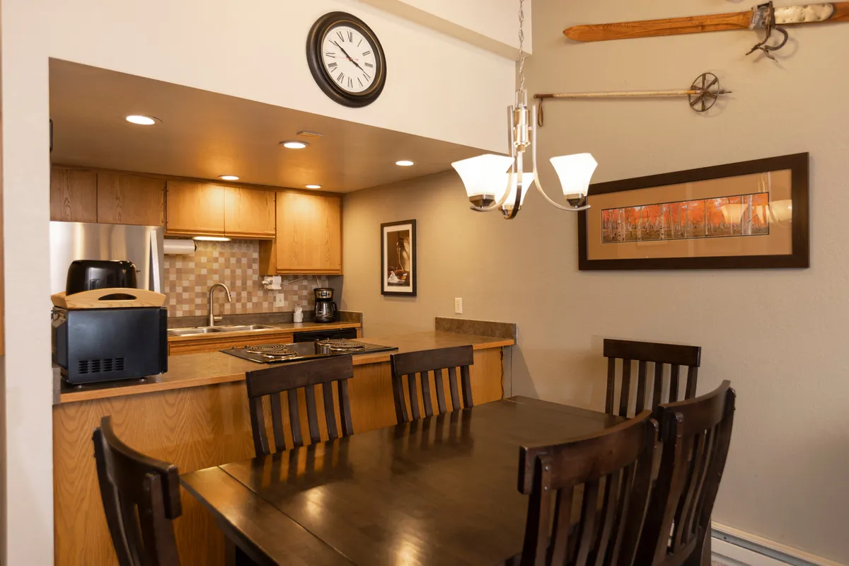A warm, inviting dining area is showcased, with a spacious table set up for group gatherings. The adjacent kitchen counter and elegant chandelier reinforce the sense of a shared, lively space. Subtle decor like wall art and a prominent clock create a relaxed yet homey atmosphere ideal for socializing and storytelling.