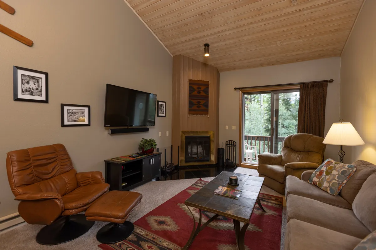 The image showcases a cozy living room interior featuring a crackling fireplace, plush sofa, and a large picture window framing breathtaking mountain views. The inviting space is perfect for relaxing with a warm drink, entertaining friends, or watching the sunset after a day on the slopes.