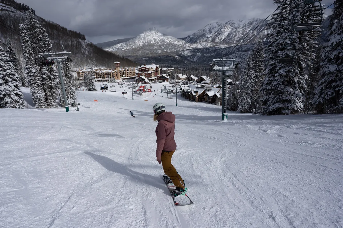 A dynamic winter scene at Purgatory Resort showcasing a snowboarder in action, ski resort buildings nestled among pine trees, ski lifts ascending the mountain, and a dramatic snow-capped mountain backdrop. The atmosphere buzzes with the excitement of winter sports and après-ski activities.