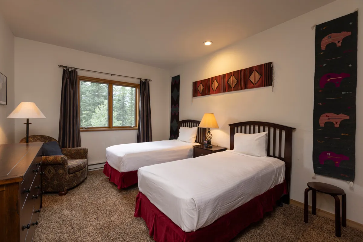 A cozy bedroom with twin beds, a window offering a pleasant view and natural light, a decorative wall hanging, and a warm lamp. The setup suggests flexible sleeping arrangements suitable for kids or guests, all in an inviting and restful setting.